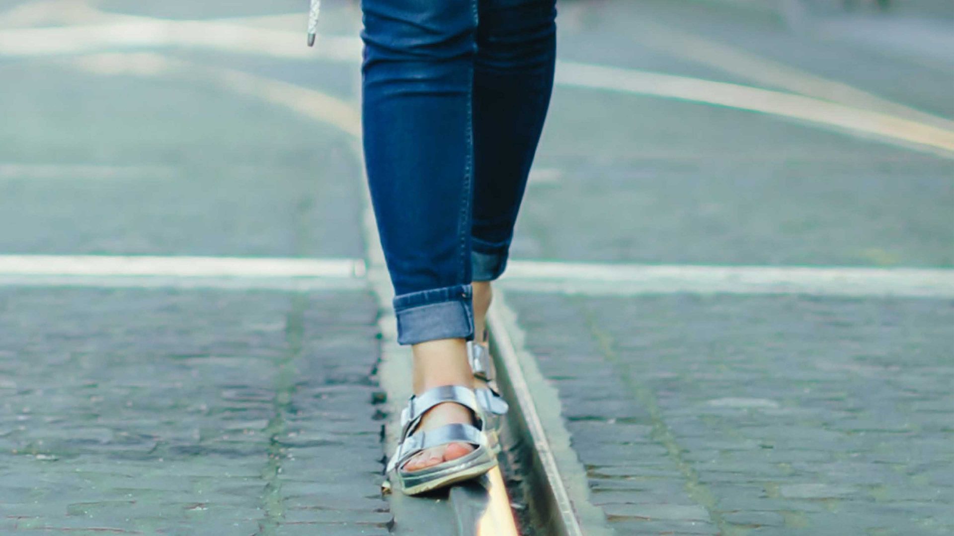Eine Frau läuft in silbernen Hausschuhen und blauer Jeans auf einem Straßenbahngleis.