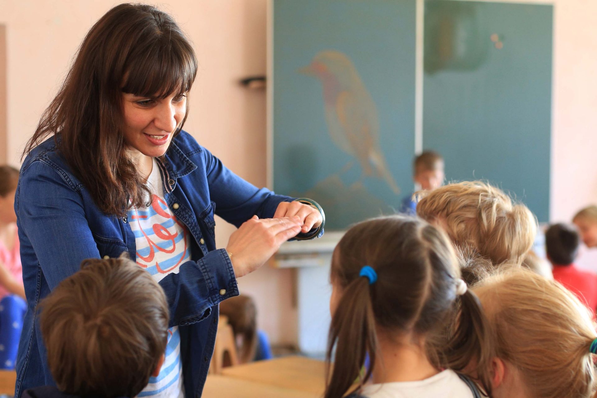 Eine Lehrerin spricht mit Kindern in einem Klassenzimmer. Im Hintergrund befindet sich eine Tafel an der Wand.