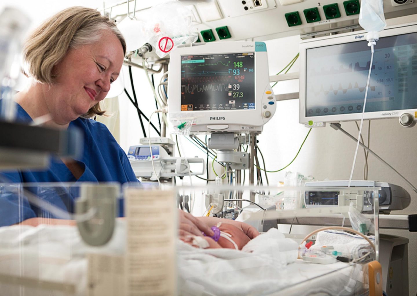 Eine Krankenschwester mittleren alters lächelt ein Kind auf der Geburtenstation eines Krankenhauses an. Im Hintergrund sind Überwachsungsinstrumente zu sehen.